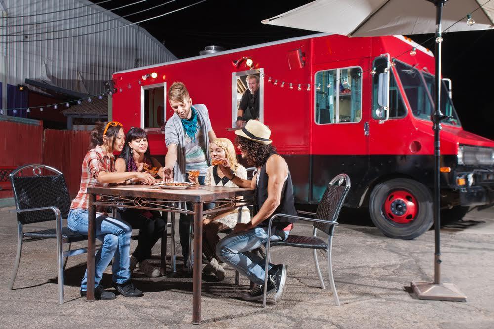 Group of five young adults enjoying a meal at an outdoor table near a red food truck, with a street lamp illuminating the area at night.