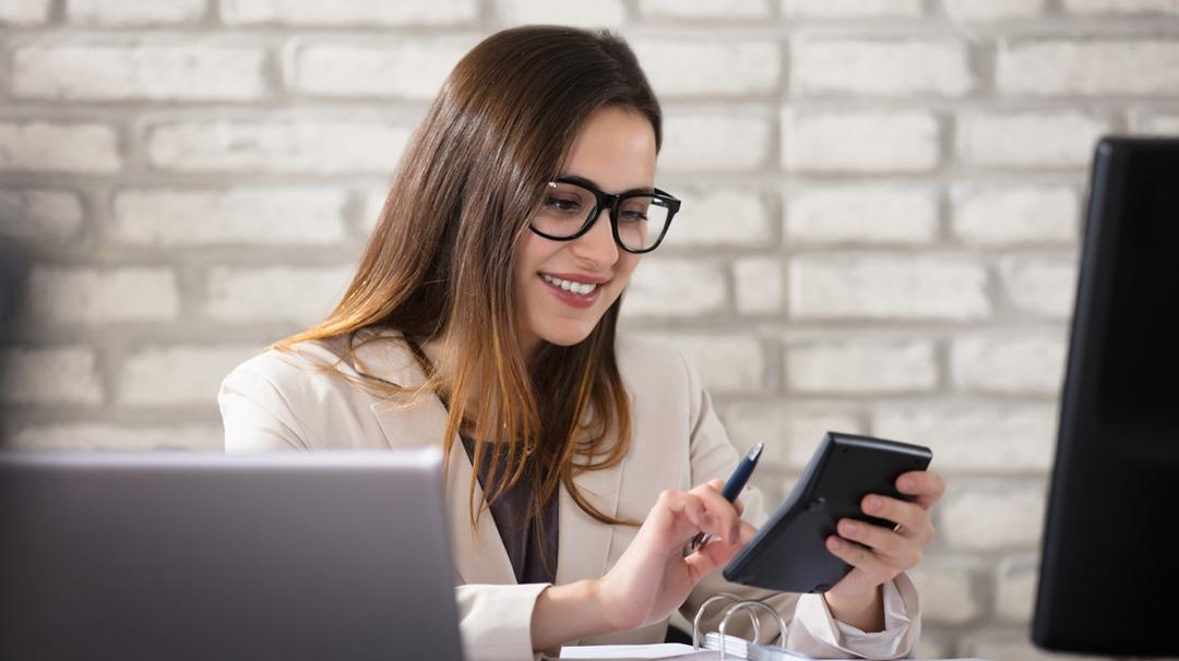 A young woman in a white blazer and glasses smiles while using a smartphone and holding a pen, sitting at a desk with multiple computer screens.