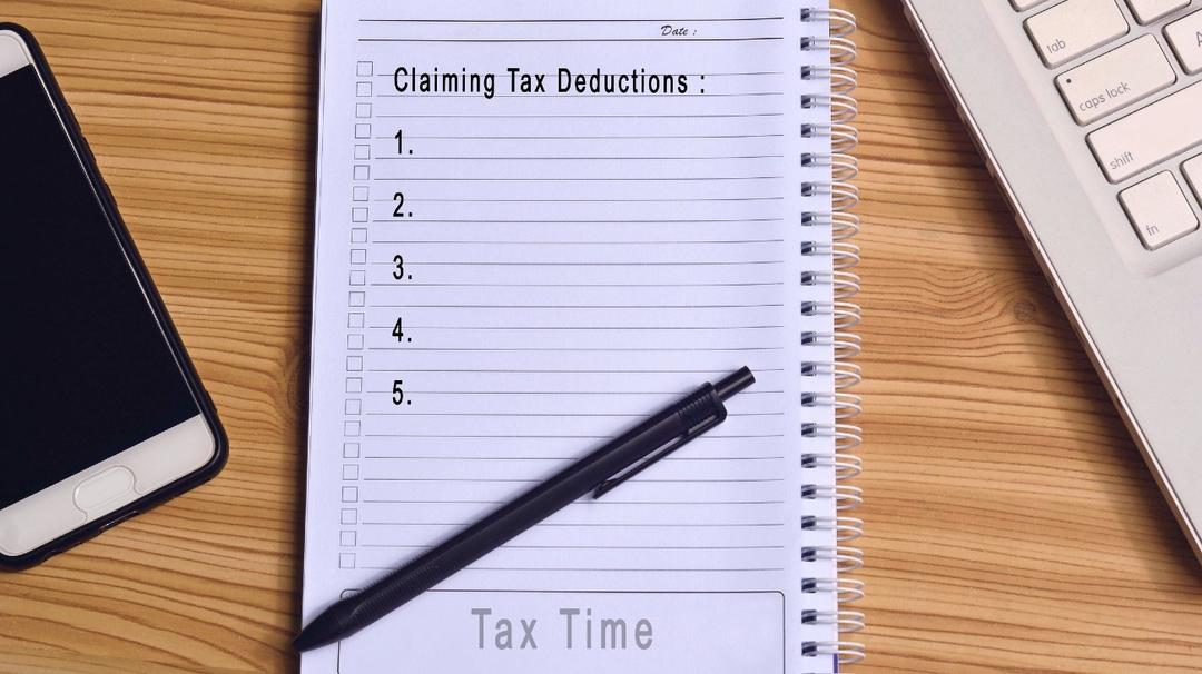 Notepad with the title "claiming tax deductions" displaying a list on a wooden desk, alongside a pen, smartphone, and keyboard.