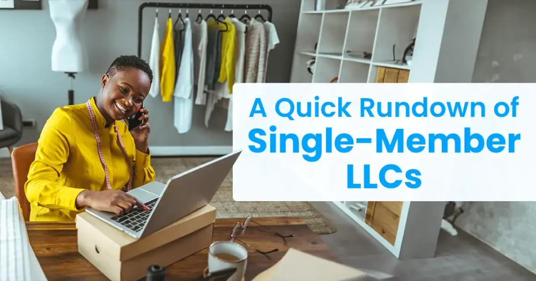 A smiling woman in a yellow blouse talks on the phone while using a laptop, sitting at a desk with a sign titled "a quick rundown of single-member llcs.